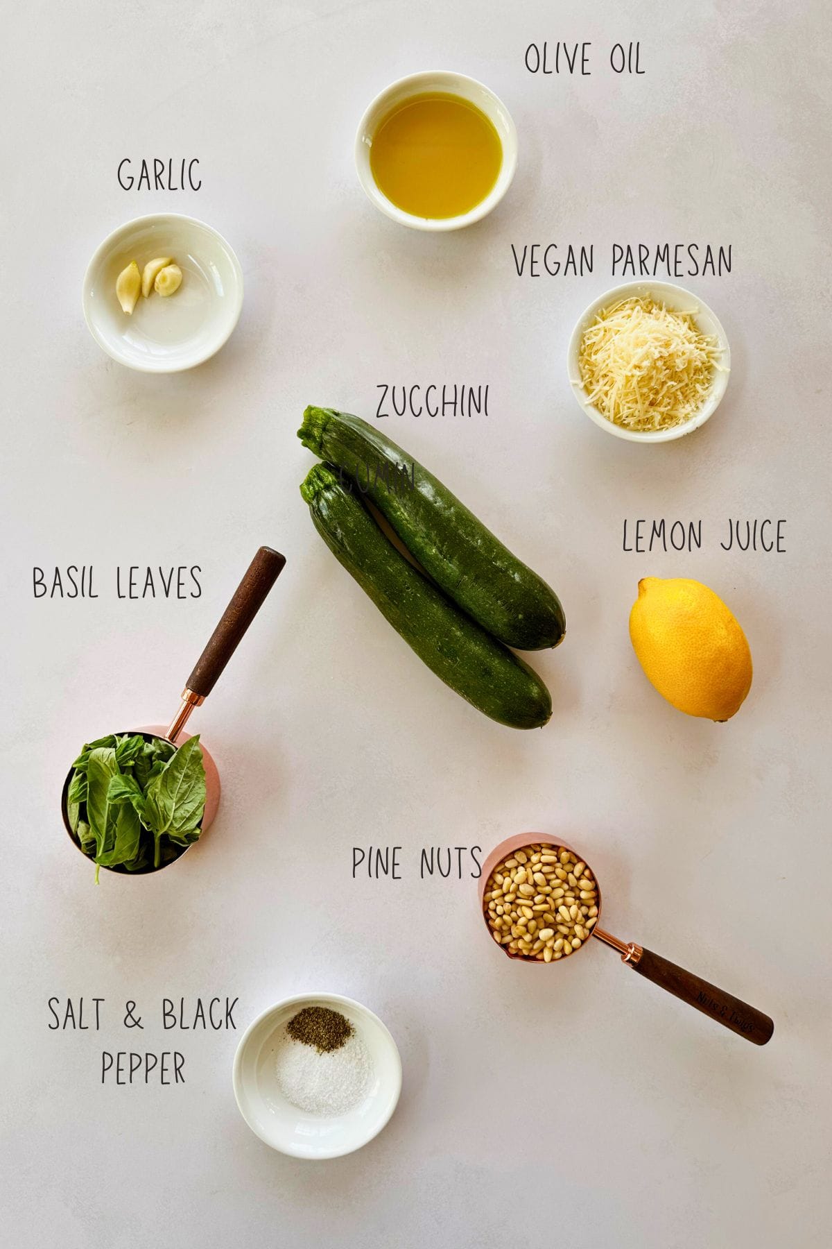 Flat lay of ingredients for zucchini pesto, including two zucchinis, fresh basil leaves, garlic cloves, vegan parmesan, lemon, olive oil, pine nuts, salt, and black pepper—all labeled