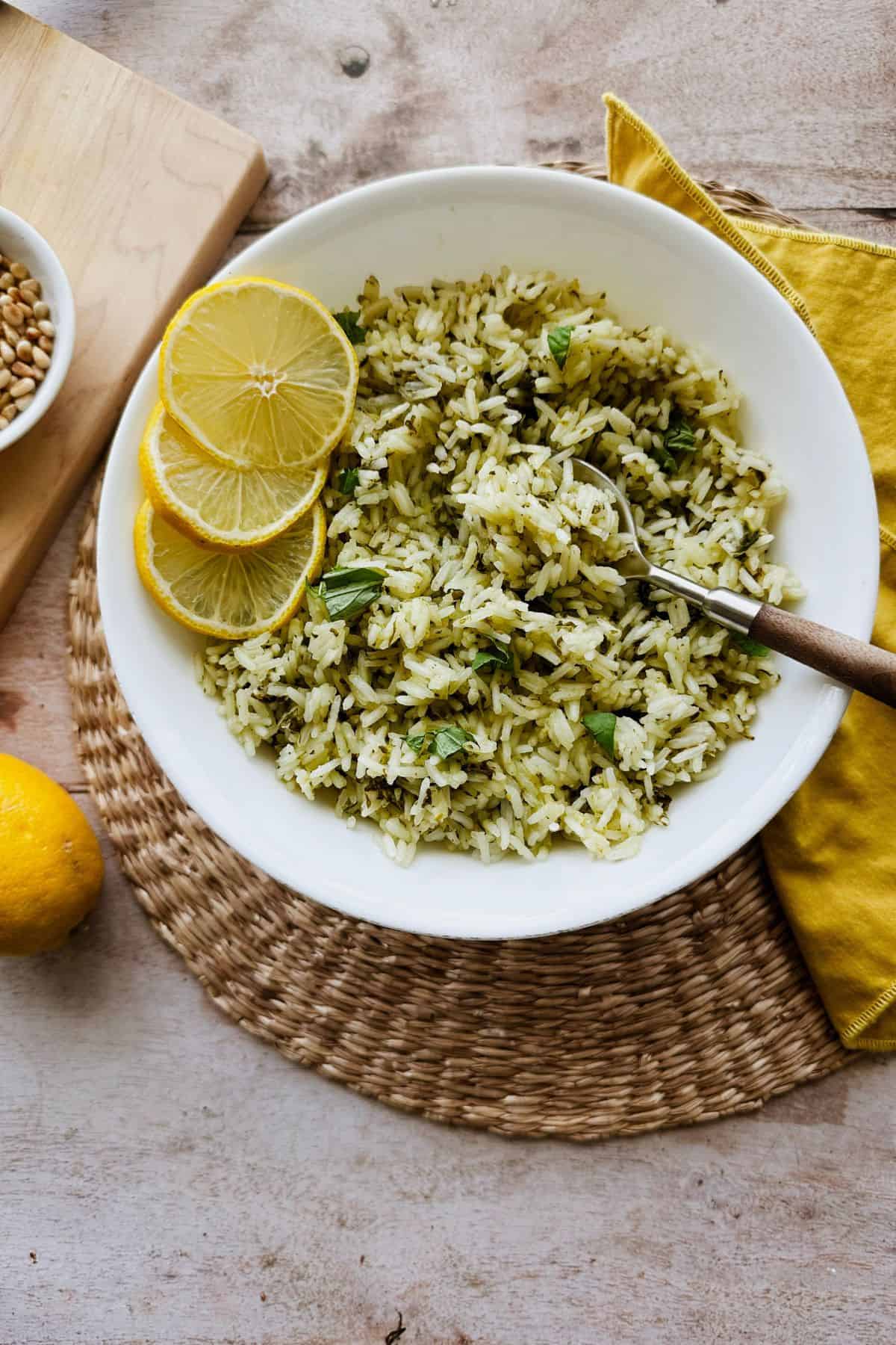 Overhead view of basil rice in a white bowl, garnished with lemon slices and fresh basil, with a spoon tucked in and surrounded by a warm-toned tablescape with a wicker placemat and yellow napkin.