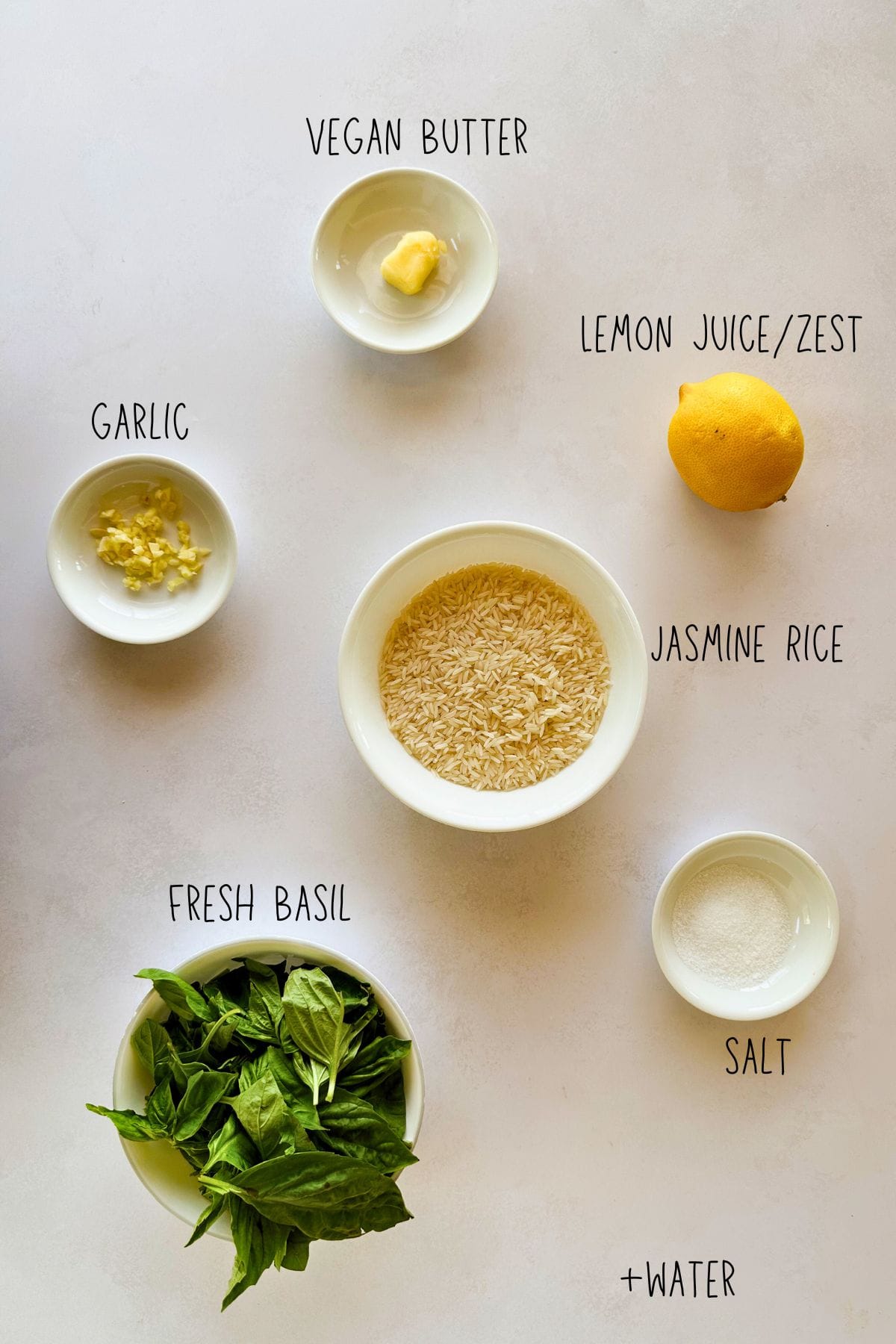 Overhead shot of ingredients for basil rice, including jasmine rice, fresh basil, lemon, garlic, vegan butter, salt, and water, each labeled in white bowls on a light background.