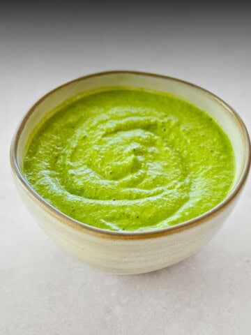 Close-up of a rustic ceramic bowl filled with creamy green basil sauce, swirled neatly on top. The sauce has a smooth, velvety texture and vibrant green color, set against a light grey background.