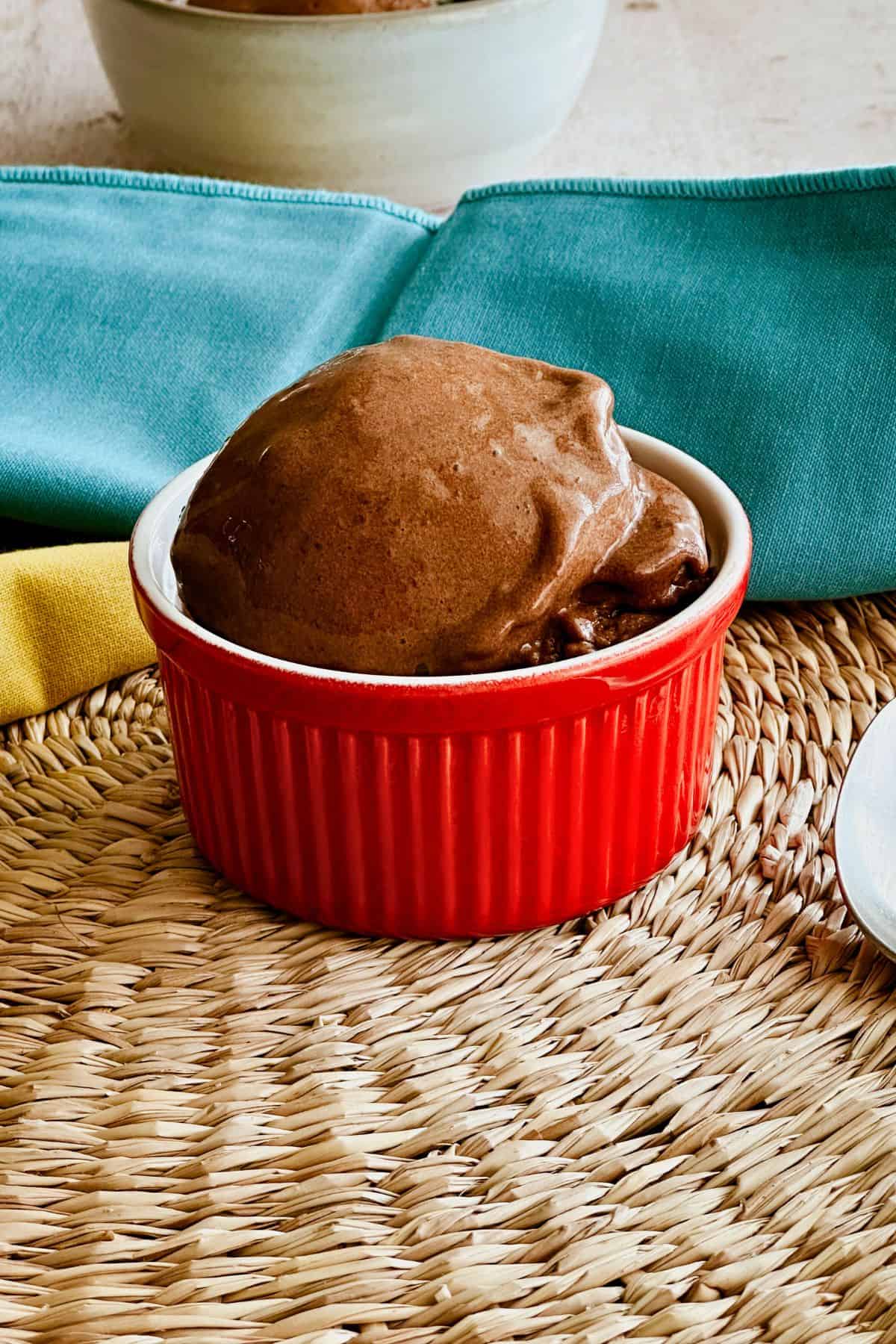 Close-up of creamy chocolate banana ice cream served in a small red ramekin, placed on a woven placemat with a teal napkin and yellow cloth in the background.