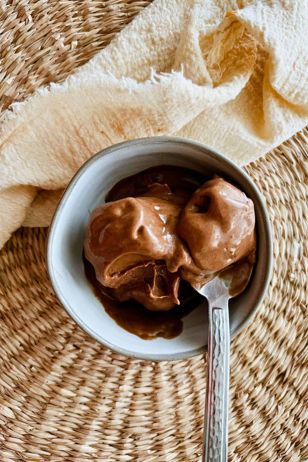 Overhead view of creamy chocolate banana ice cream served in a light blue bowl with a silver spoon, placed on a round woven placemat with a cream-colored textured cloth beside it.