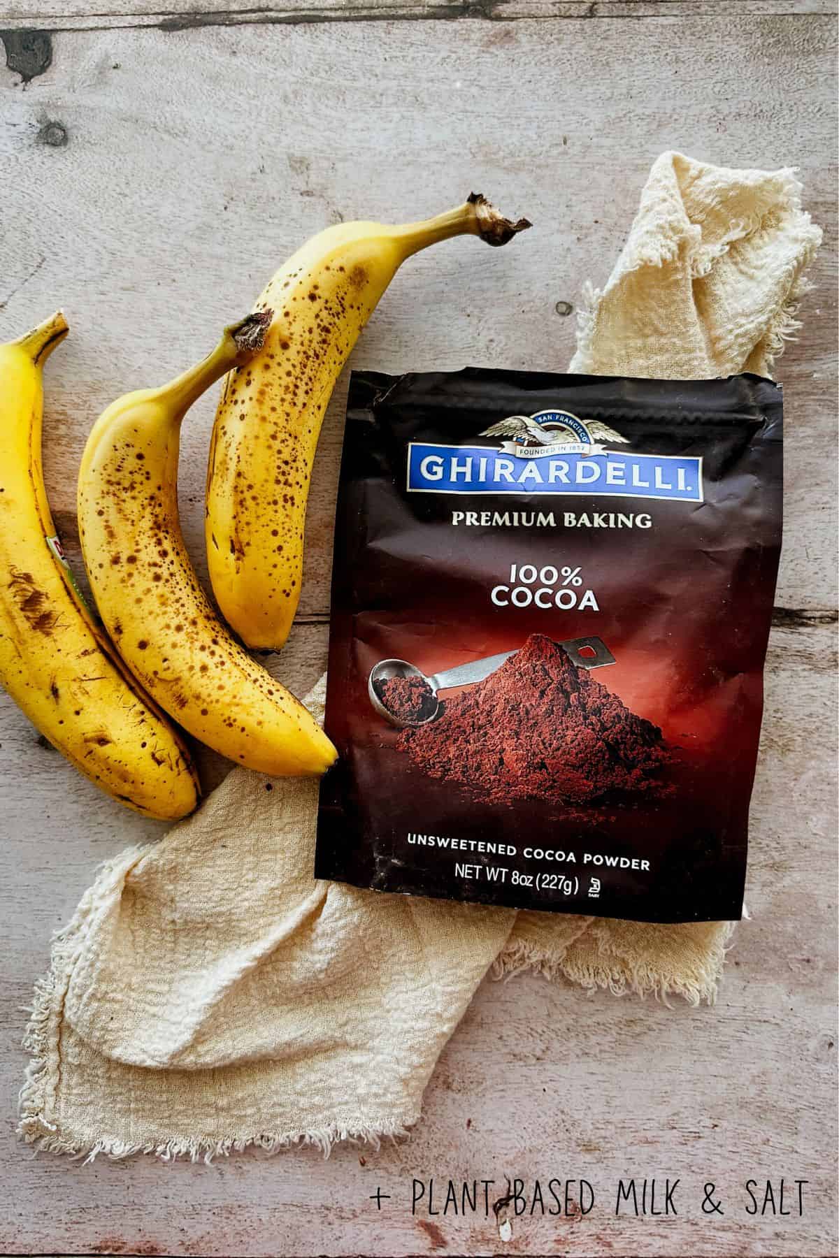 Overhead shot of three ripe bananas with brown spots arranged next to a bag of Ghirardelli Premium Baking 100% unsweetened cocoa powder, all set on a light wooden surface with a cream-colored textured cloth. Text in the bottom right corner reads 