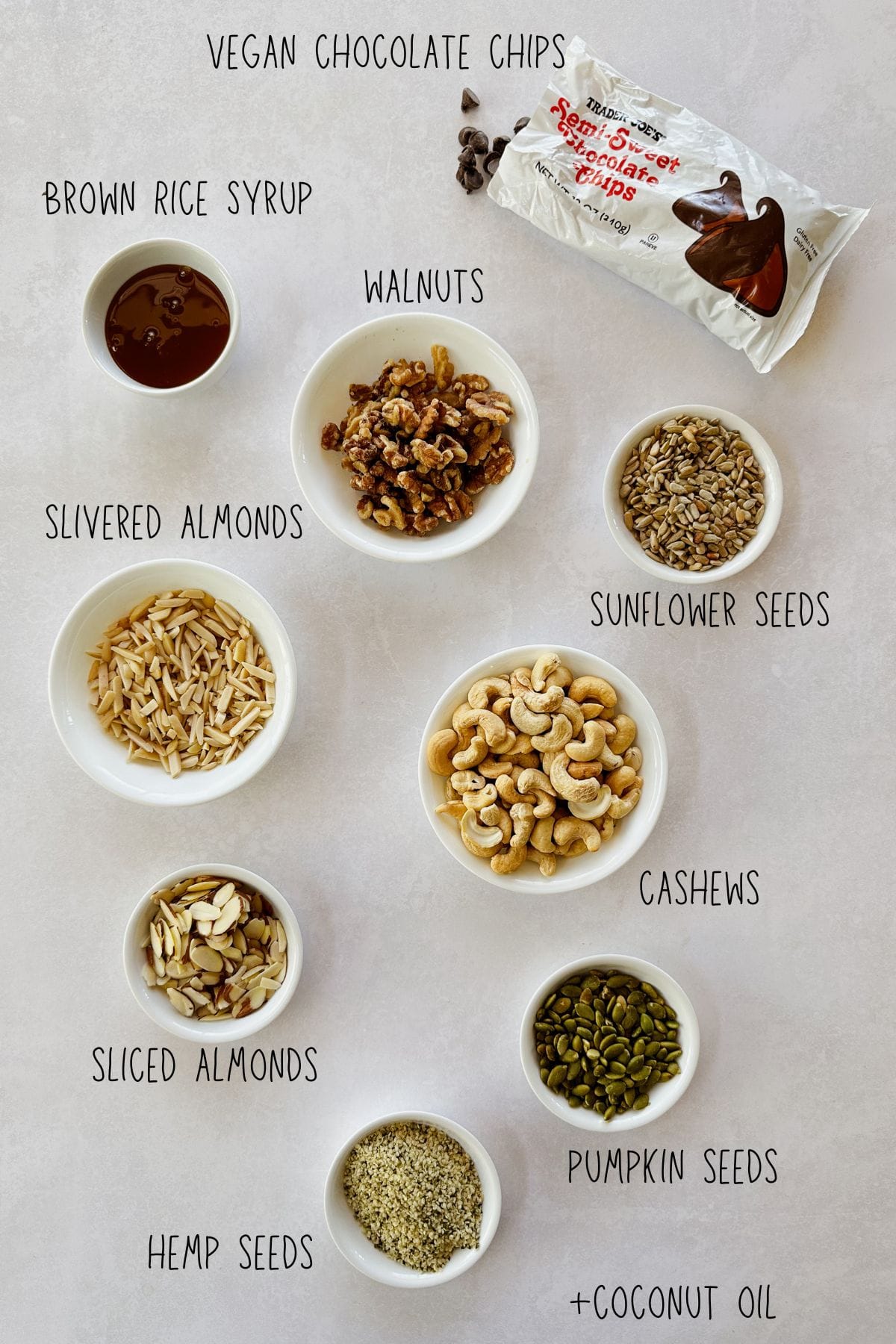 Overhead view of small bowls filled with ingredients for vegan chocolate bark: vegan chocolate chips, brown rice syrup, walnuts, sunflower seeds, cashews, slivered almonds, sliced almonds, pumpkin seeds, coconut oil, and hemp seeds, all labeled.