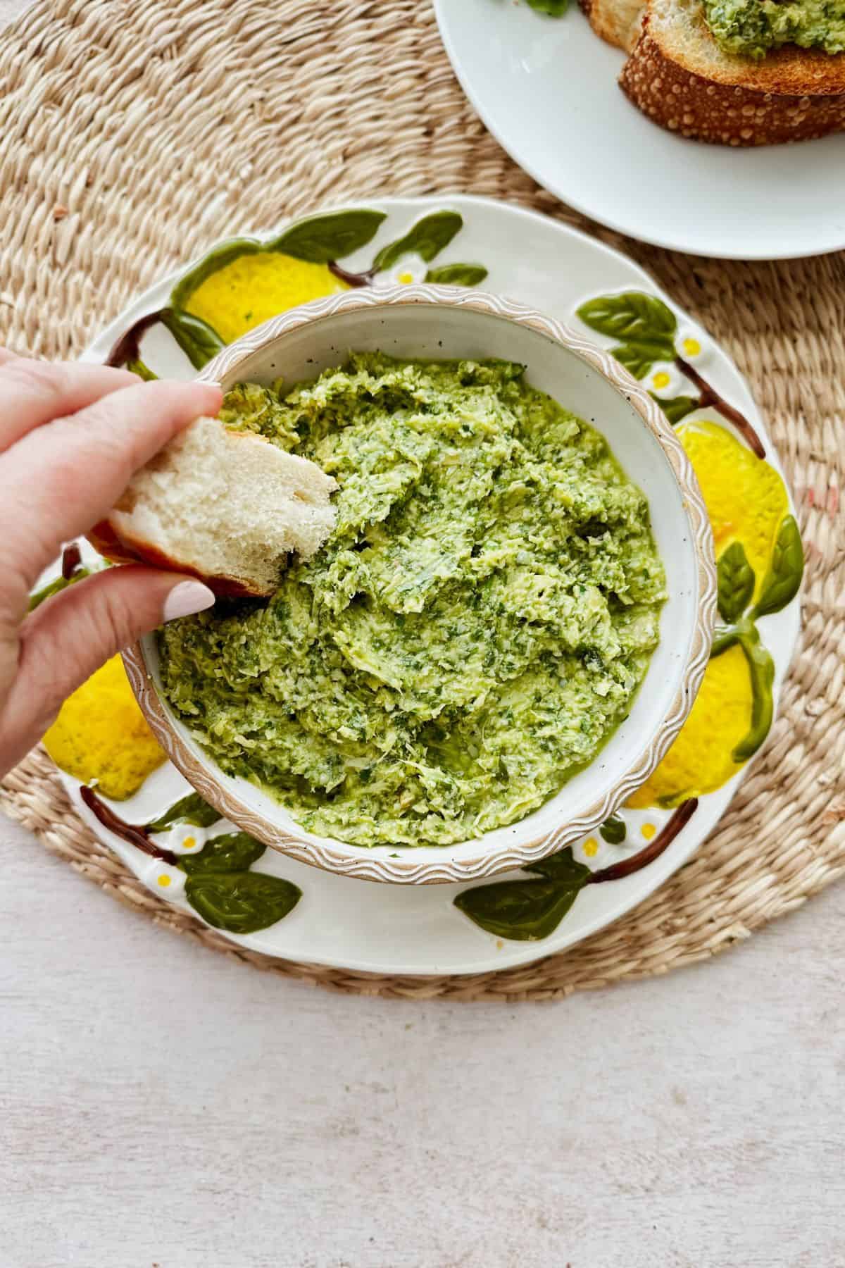 Hand dipping crusty bread into a bowl of lemony arugula artichoke dip, a fresh dairy-free and nut-free spread full of zesty herbs and bright flavor.