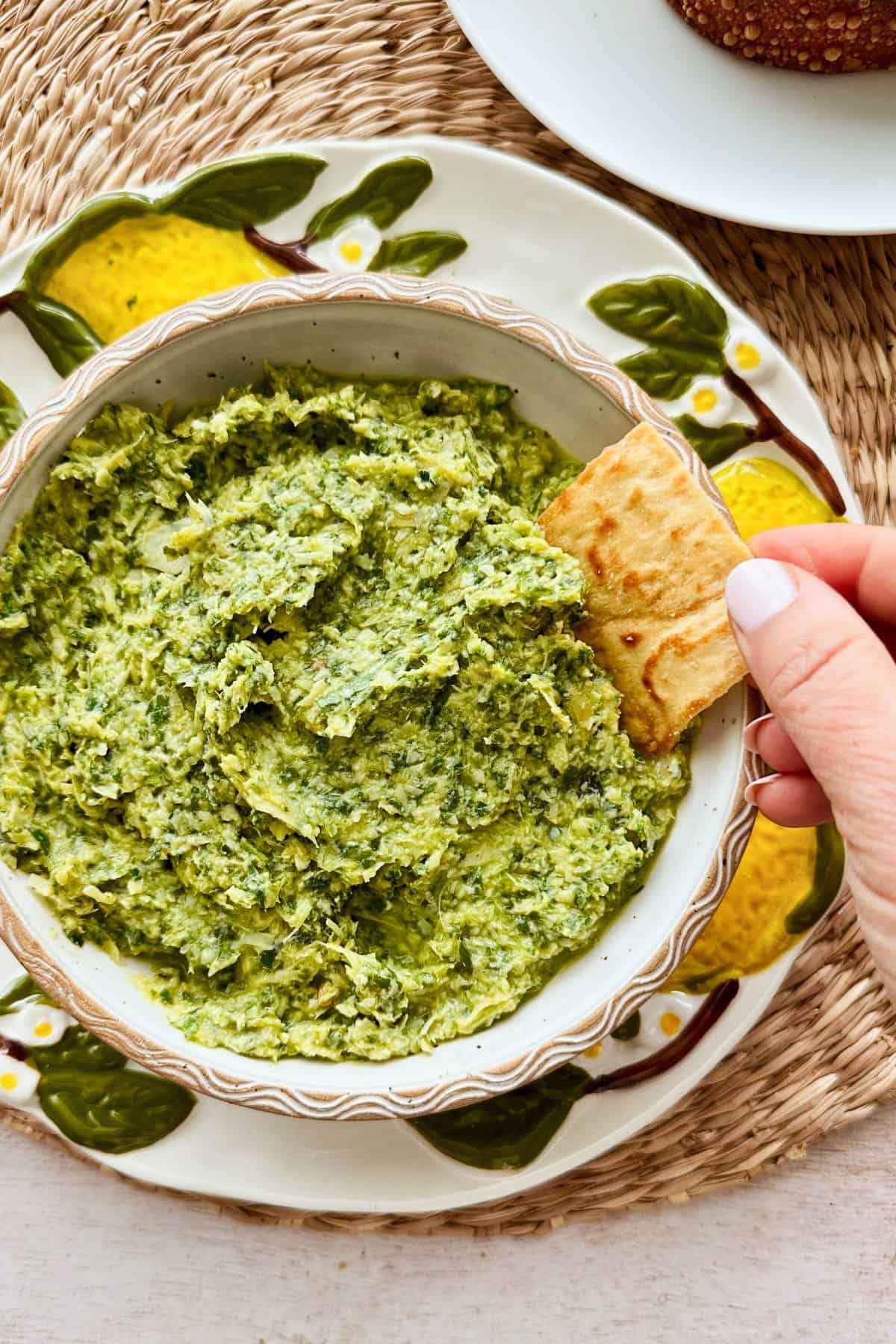 Hand dipping a pita chip into creamy lemony arugula artichoke dip, a healthy vegan and dairy-free appetizer with fresh herbs and citrus.