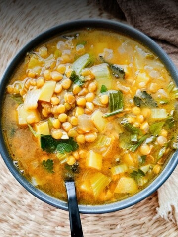 A top-down view of a bowl of lemony lentil soup filled with tender potatoes, green leafy vegetables, and brown lentils, served with a black spoon resting in the bowl on a woven placemat. A beige napkin and brown cloth are partially visible, adding warmth and texture to the scene.