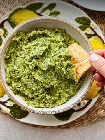 Hand dipping a pita chip into creamy lemony arugula artichoke dip. Bowl is sitting on top of a decorative plate.