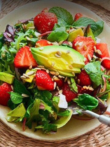 A fresh strawberry avocado salad on a beige plate, featuring vibrant greens, sliced avocado, halved strawberries, fresh mint leaves, sunflower seeds, and pecans, set on a woven placemat with whole strawberries and a light cloth in the background.