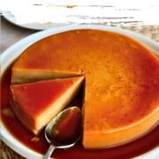 A slice of vegan flan topped with golden caramel sauce on a white plate, with the rest of the flan and coffee in the background. Text on the graphic says, "the best vegan flan" and "This flan will fool everyone."