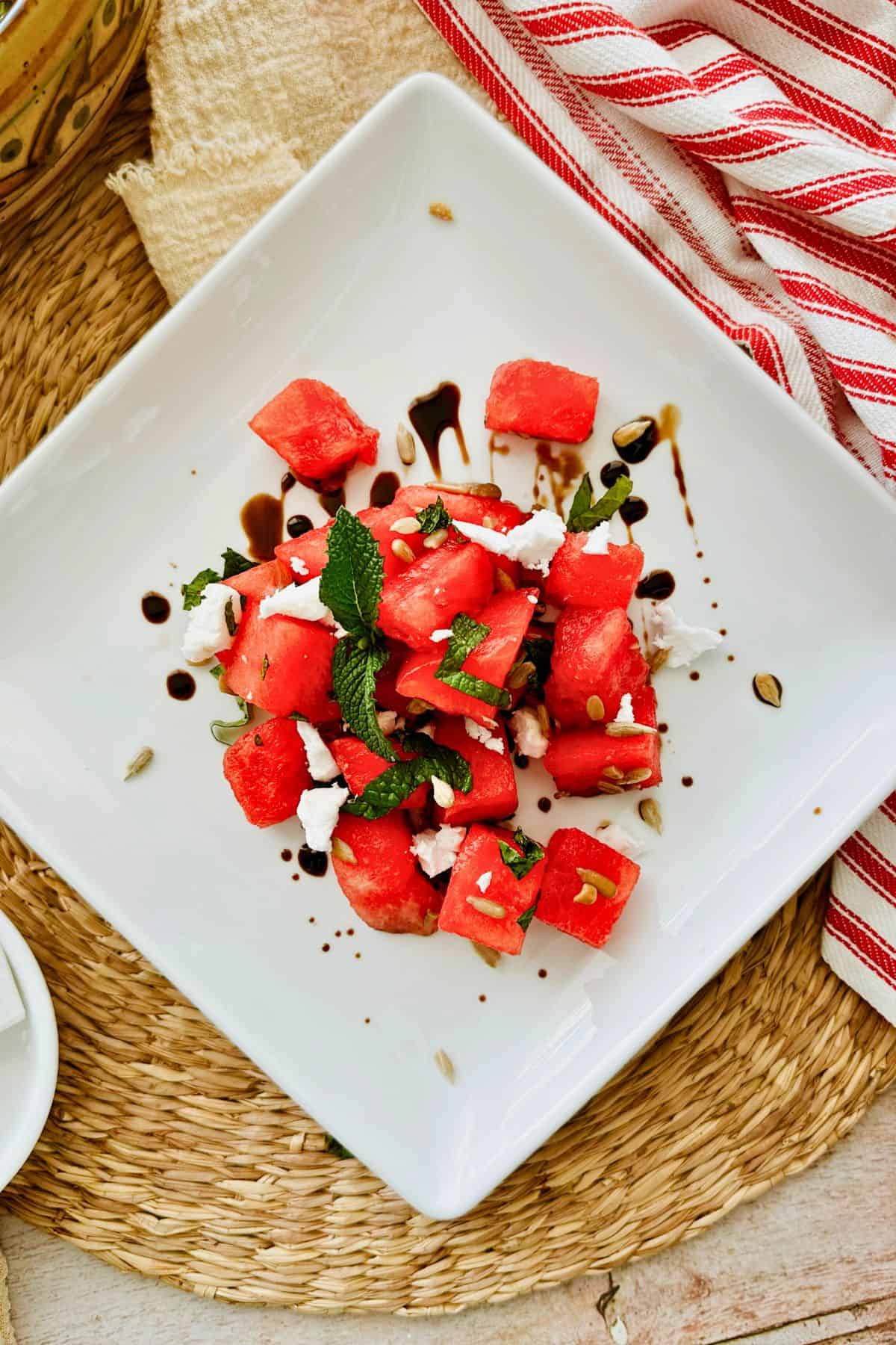Vegan watermelon salad with fresh mint, vegan feta, sunflower seeds, and balsamic glaze on a white square plate.