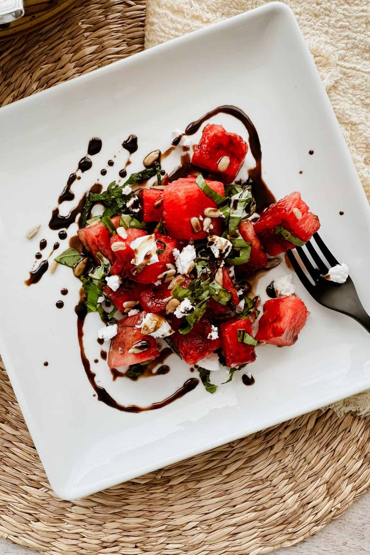 Vegan watermelon basil salad topped with vegan feta, sunflower seeds, and fresh herbs, drizzled with balsamic glaze and served on a white square plate with a black fork.