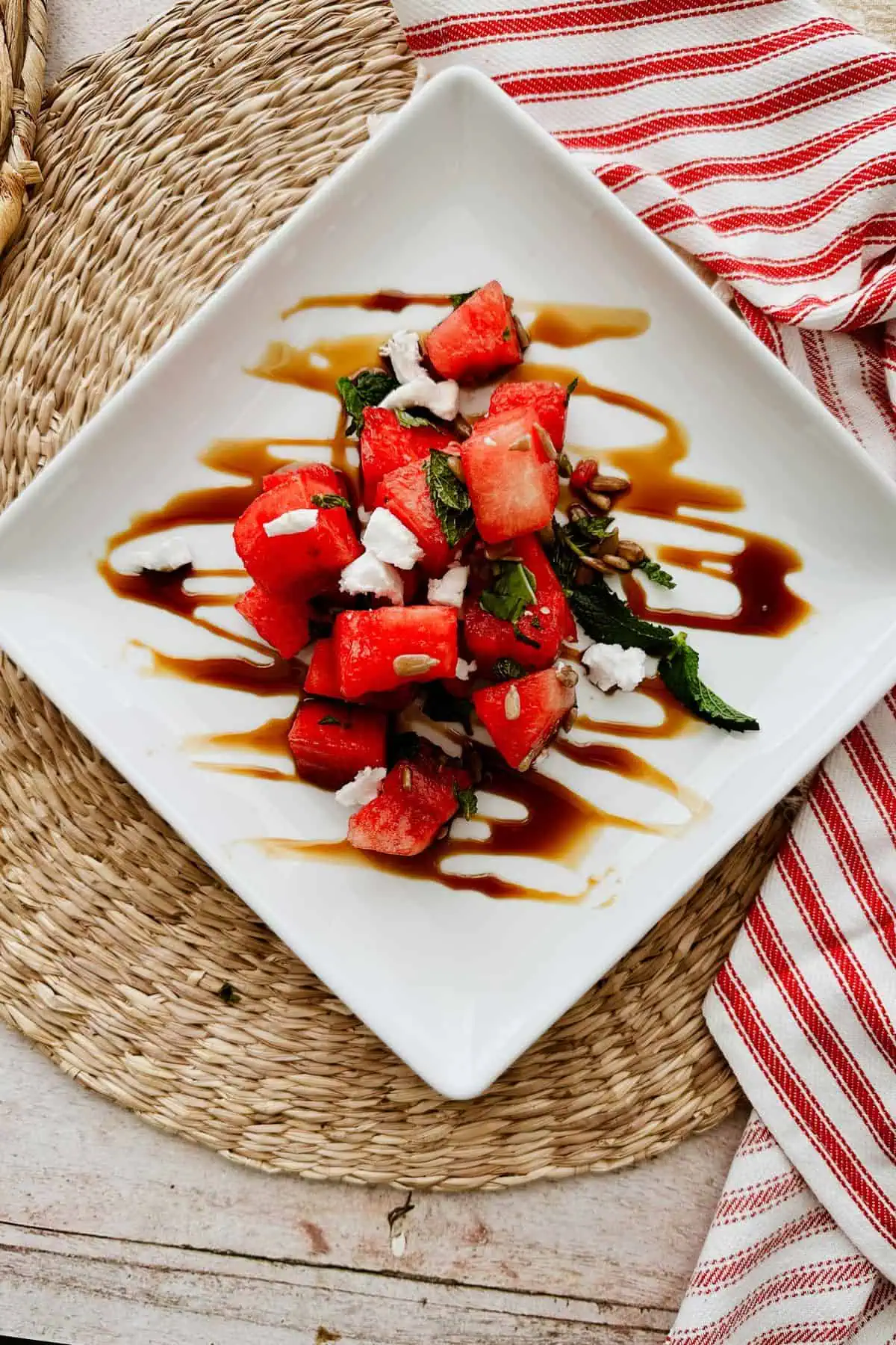 Vegan watermelon salad with vegan feta, sunflower seeds, mint, and basil, served on a white square plate with a balsamic glaze drizzle and a red-striped napkin on the side.