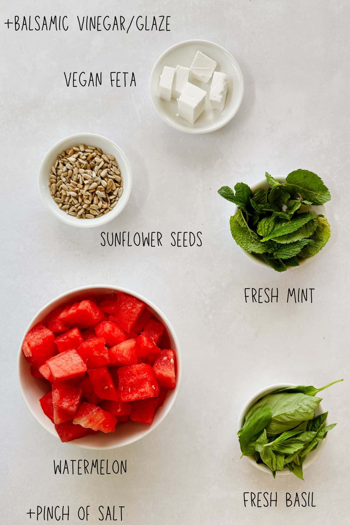Bowls of chopped watermelon, vegan feta, sunflower seeds, fresh mint, and basil arranged on a white surface with labels, ingredients for vegan watermelon basil salad.