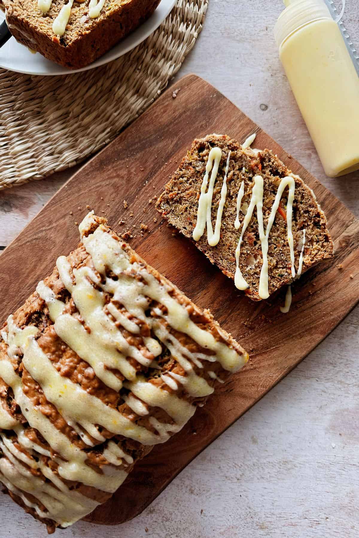 A sliced loaf of zucchini carrot apple bread on a wooden cutting board, drizzled with cream cheese orange glaze. One slice is separated and decorated with extra glaze. A squeeze bottle of glaze is visible nearby. The loaf has a moist, dense texture with visible bits of carrot and zucchini.