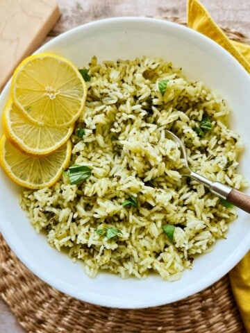 Bowl of fluffy jasmine rice mixed with chopped fresh basil and garnished with lemon slices, served with a spoon on a woven placemat and yellow napkin.