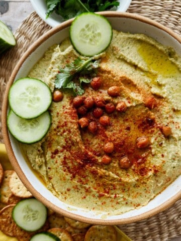 A large bowl of creamy cucumber hummus topped with paprika-dusted roasted chickpeas, fresh cucumber slices, and a drizzle of olive oil, surrounded by crackers, cucumber slices, a lemon, and fresh herbs on a woven placemat