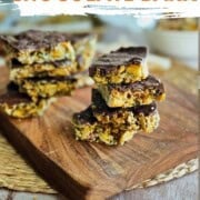 Close-up of stacked squares of vegan chocolate bark on a wooden cutting board. The bark has a golden, nutty base and a thick layer of glossy dark chocolate on top. The texture is crunchy, with visible seeds and nuts, and the chocolate is finished with a slight shine. Text on the image reads “Vegan Chocolate Bark – So Easy!” with the website nutsandtwigs.com at the bottom.