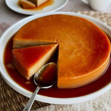 Close-up of a vegan flan with a slice removed, a spoon resting in the caramel sauce, and a blurred plate of sliced flan in the background.