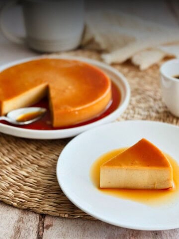 A whole vegan flan with a golden caramel top, one slice removed and served on a white plate in the foreground, with coffee cups in the background on a woven placemat.