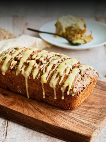 Loaf of zucchini, carrot, and apple bread drizzled with orange cream cheese glaze, resting on a wooden cutting board. A slice is served on a plate in the background with a fork.