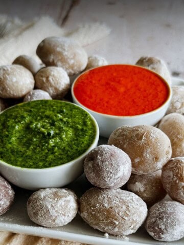 Close-up of Canarian papas arrugadas, small wrinkled potatoes covered in a white salty crust, arranged around two bowls of sauce. One bowl contains bright green mojo verde, and the other holds vivid red mojo rojo