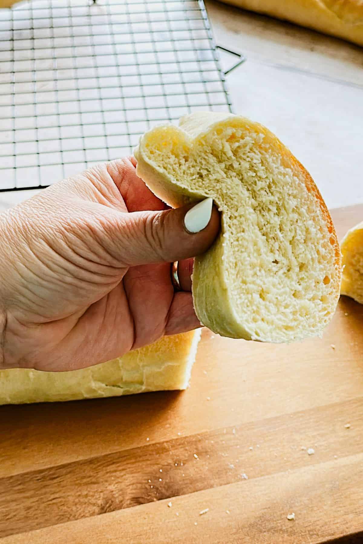 Close-up of a hand holding a slice of freshly baked Cuban bread, showing its soft, fluffy crumb and thin golden crust, with a cooling rack and loaf in the background.