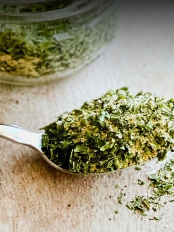 A spoonful of homemade ranch dressing mix with dried parsley, dill, chives, garlic, and onion powder, resting in front of a glass jar filled with the ranch seasoning blend. Loose flakes of the dried ranch mix are scattered on the wooden surface.