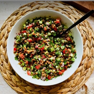 Overhead view of lentil tabbouleh salad in a white bowl with a spoon, showing fresh parsley, cucumbers, tomatoes, onions, and lentils on a woven placemat.