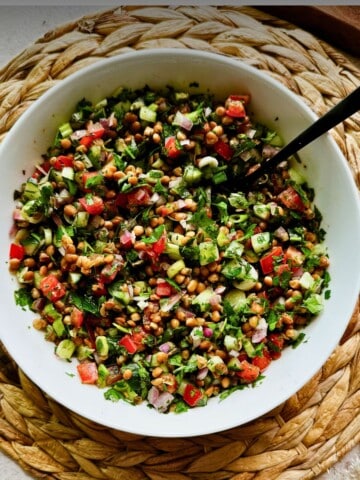 Overhead view of lentil tabbouleh salad in a white bowl with a spoon, showing fresh parsley, cucumbers, tomatoes, onions, and lentils on a woven placemat.