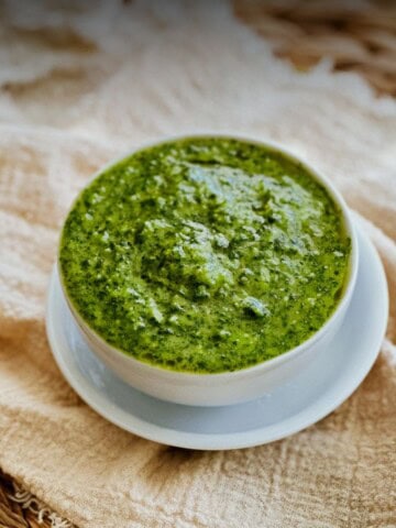 Close-up of a small white bowl filled with mojo verde, a bright green Canarian sauce made with cilantro, garlic, and olive oil. The bowl sits on a white saucer over a beige cloth with a woven mat in the background.