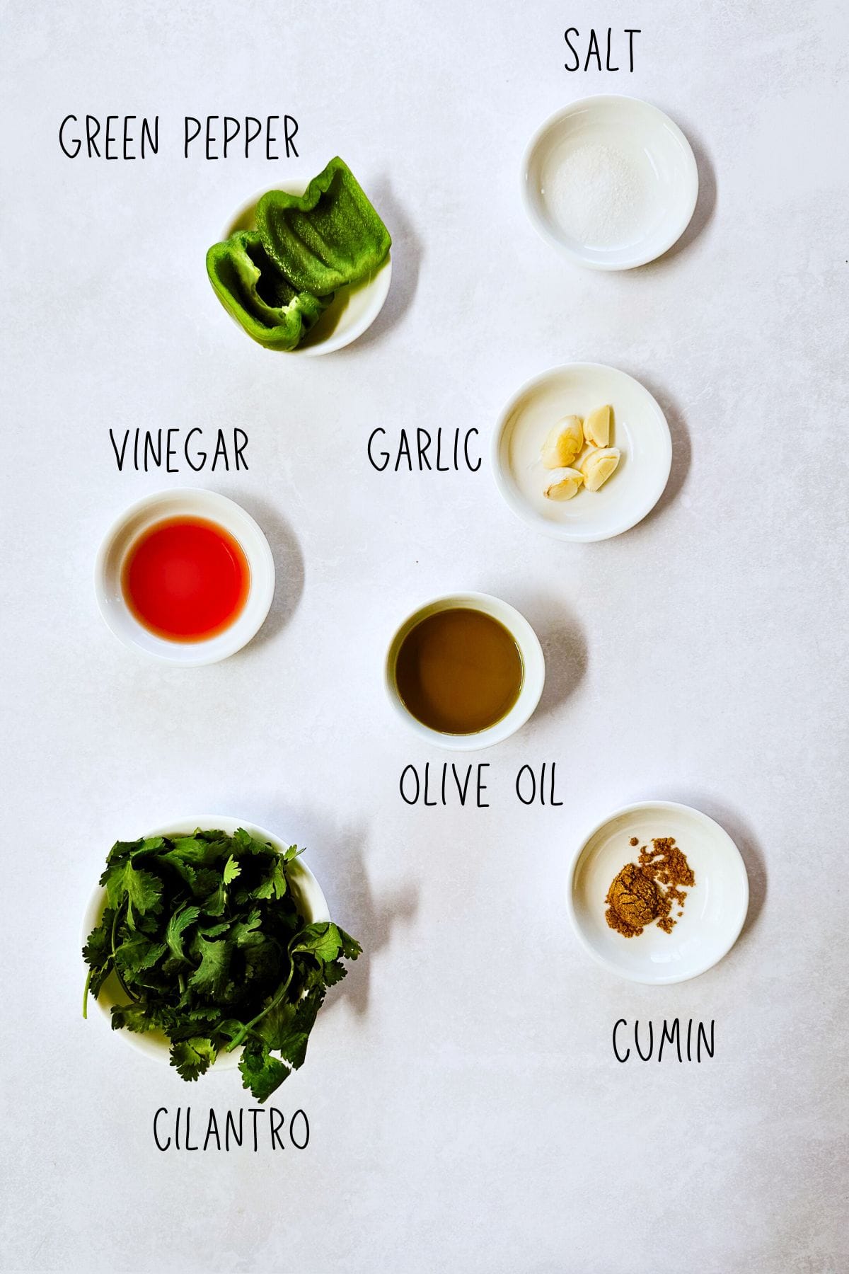 Overhead view of small white bowls arranged on a light surface, each holding ingredients for mojo verde sauce. The bowls contain green pepper, garlic cloves, olive oil, vinegar, ground cumin, and salt. A bunch of fresh cilantro is placed in a small bowl at the bottom of the image. Each ingredient is labeled with text on the photo.
