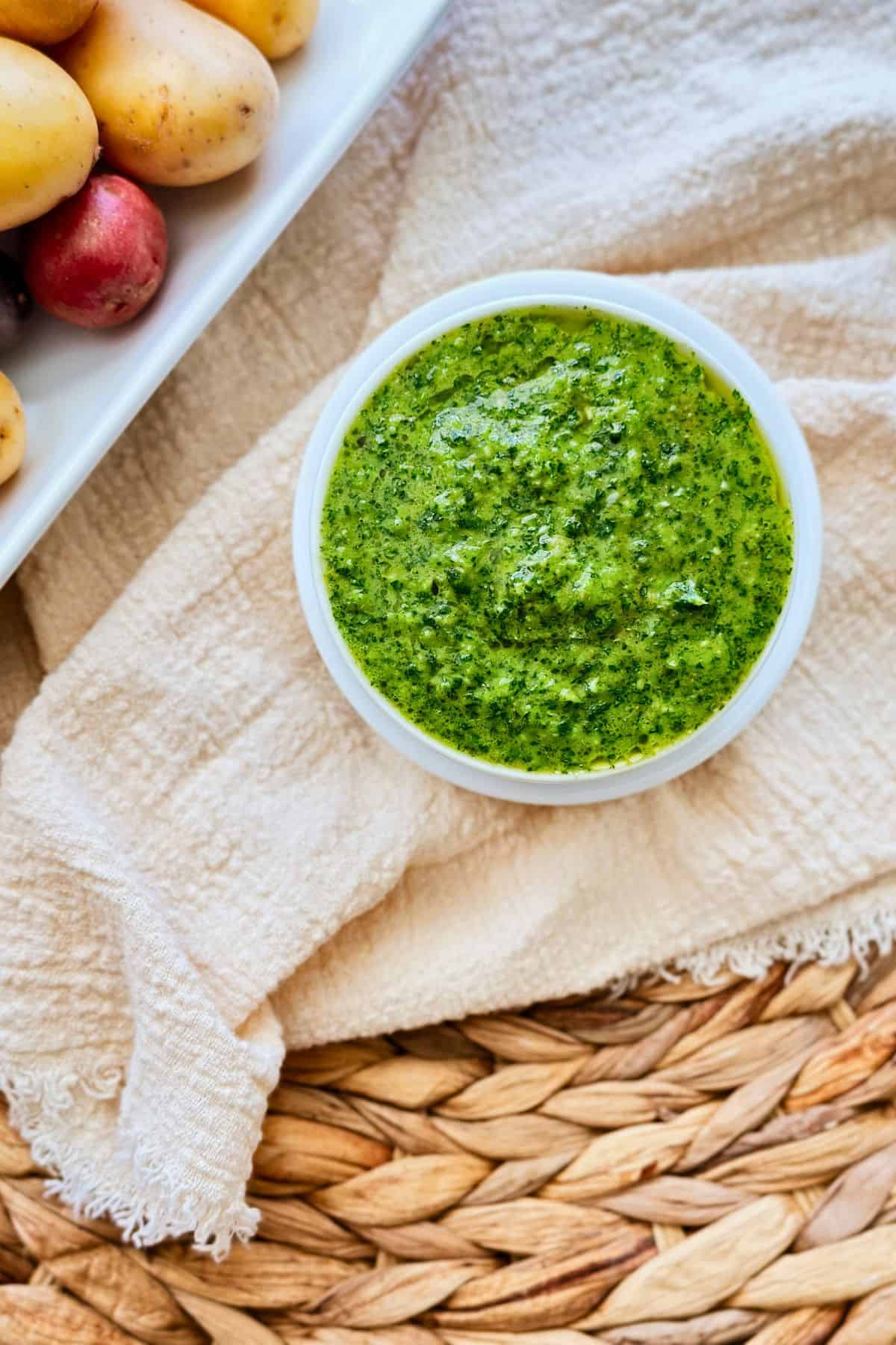 Close-up of a small white bowl filled with bright green mojo verde sauce, placed on a beige cloth and woven mat. In the corner of the image, a plate with small red and yellow potatoes is partially visible.