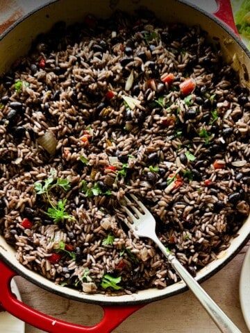 A large red Dutch oven filled with moros y cristianos, a Cuban dish of black beans and rice mixed with onions, red peppers, and garnished with fresh herbs. A fork rests inside the pot, with sides of fried plantains and salad partially visible on plates nearby.