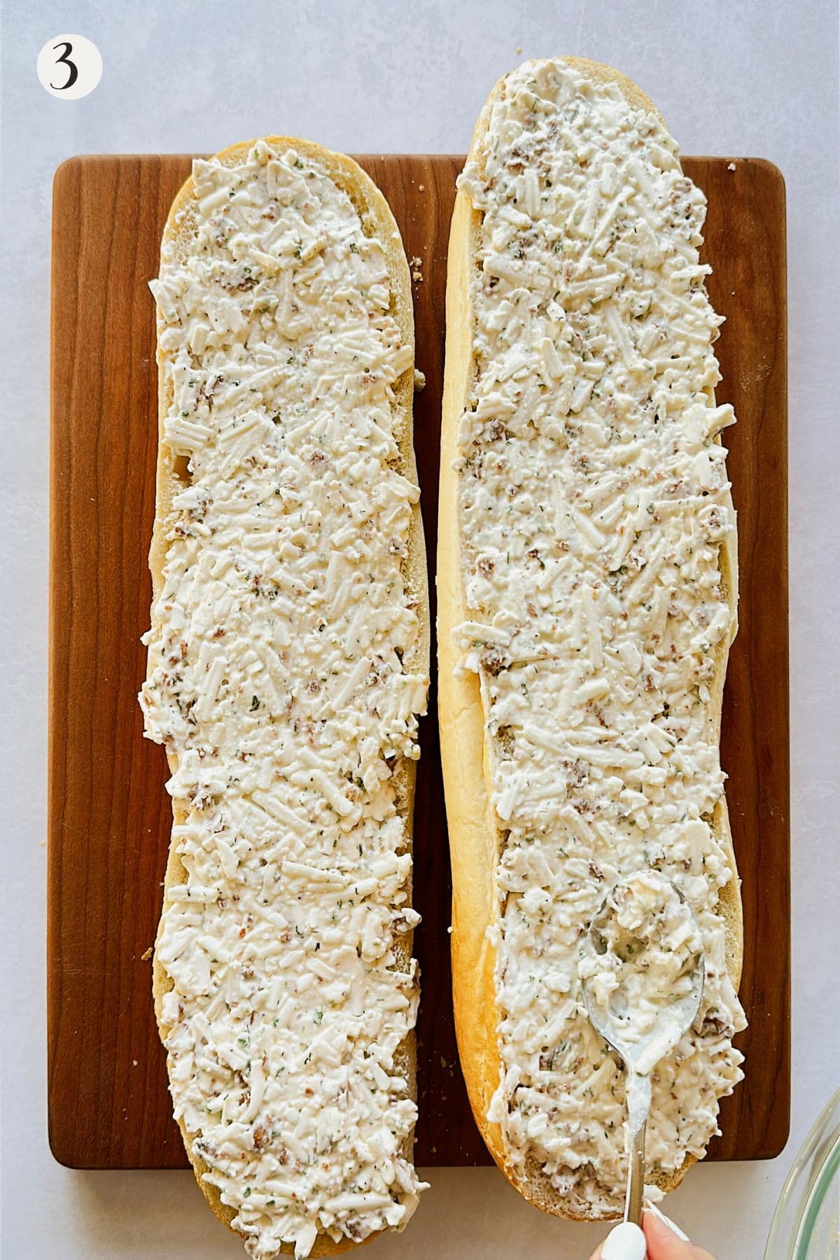 Two halves of crusty bread on a wooden board spread generously with a creamy mixture of vegan cheese, mayo, herbs, and bacon bits, ready to bake.