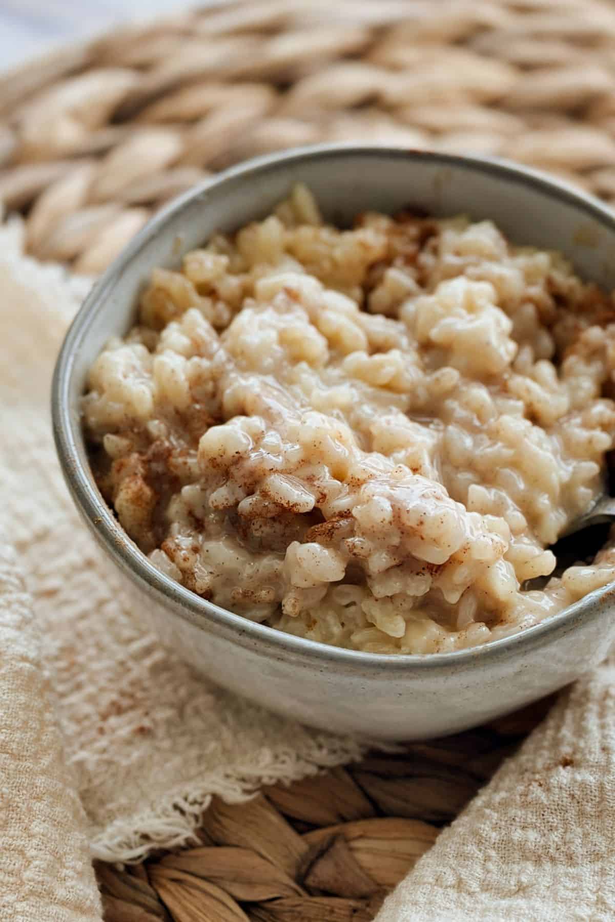 Close-up of a bowl filled with creamy vegan rice pudding. The pudding is thick and slightly glossy, with soft rice grains coated in a sweet, milky sauce and sprinkled with cinnamon. The bowl rests on a woven placemat with a light textured cloth beside it