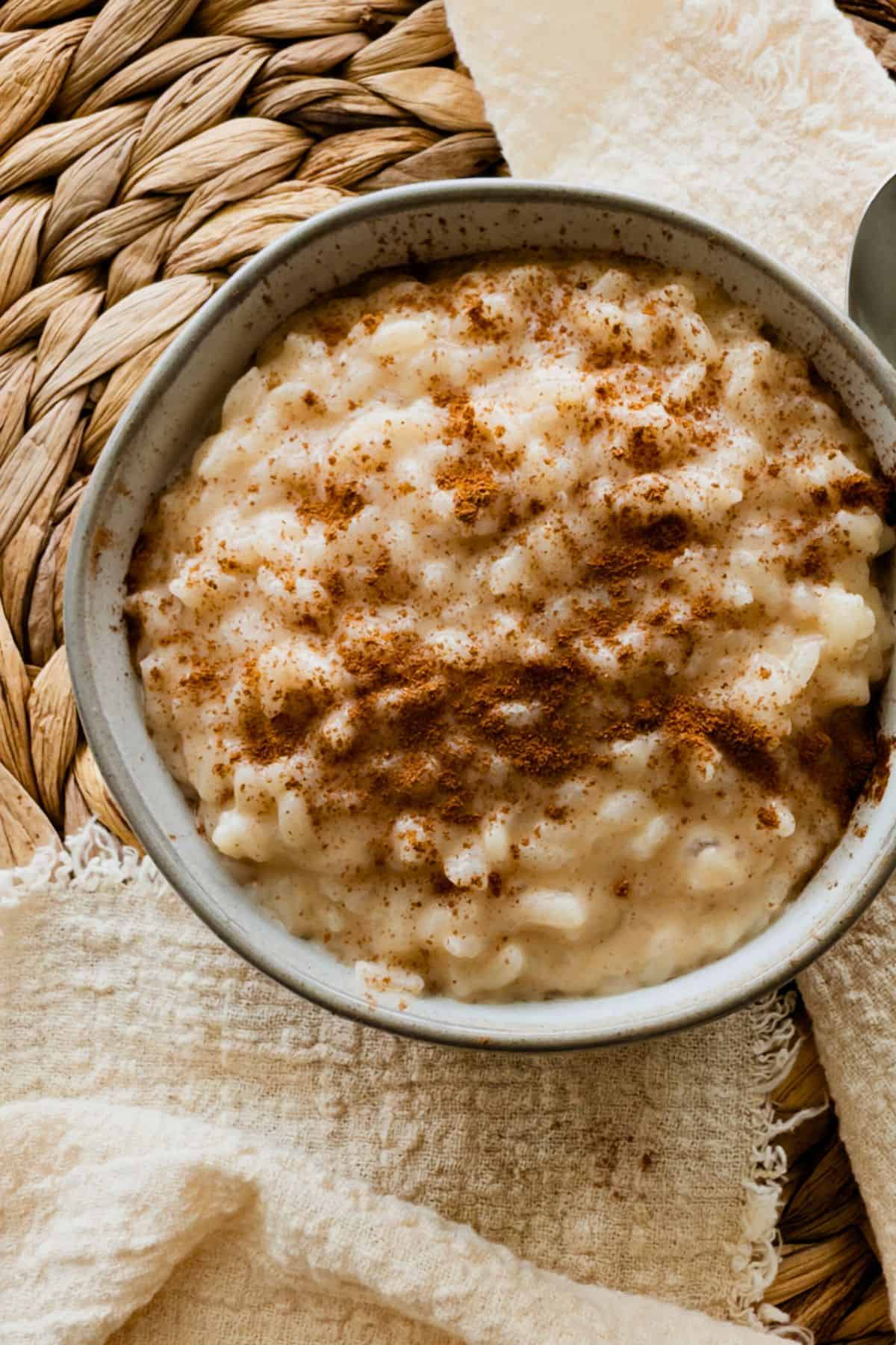 A close-up of a bowl filled with creamy vegan rice pudding. The pudding is thick and glossy, with visible tender grains of rice coated and sprinkled with ground cinnamon. There is a textured beige cloth beside it. It sits on a woven mat.