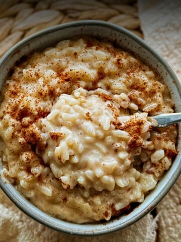 A close-up of a bowl filled with creamy vegan rice pudding. The pudding is thick and glossy, with visible tender grains of rice coated in a sauce and sprinkled with ground cinnamon. A spoon rests inside the bowl, and a textured beige cloth lies beside it.
