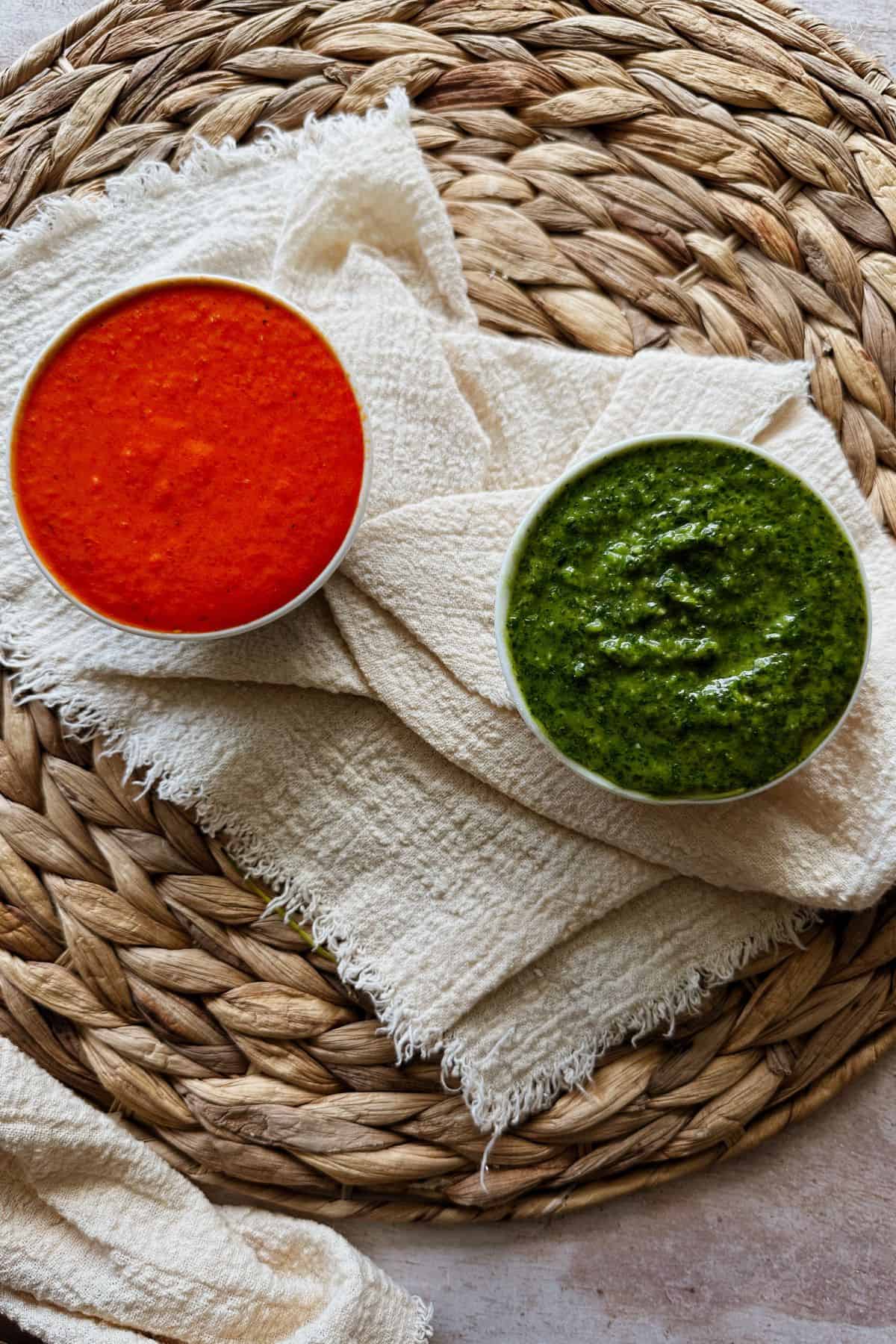 Two small bowls of sauce on a textured woven placemat with a cream-colored cloth. The bowl on the left holds a smooth, bright red sauce, mojo rojo, while the bowl on the right contains a thick green sauce, mojo verde.