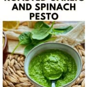 A small bowl of bright green spinach pesto with a spoon, surrounded by fresh spinach leaves and a slice of lemon. Text says, 'Winter Pesto Roasted Garlic and Spinach Pesto.'