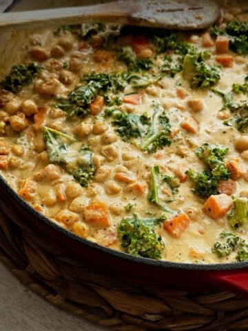 Close-up of creamy chickpea and sweet potato stew in a red pan with kale, tender chickpeas, and golden sauce. A wooden spoon rests on the edge, ready to serve.