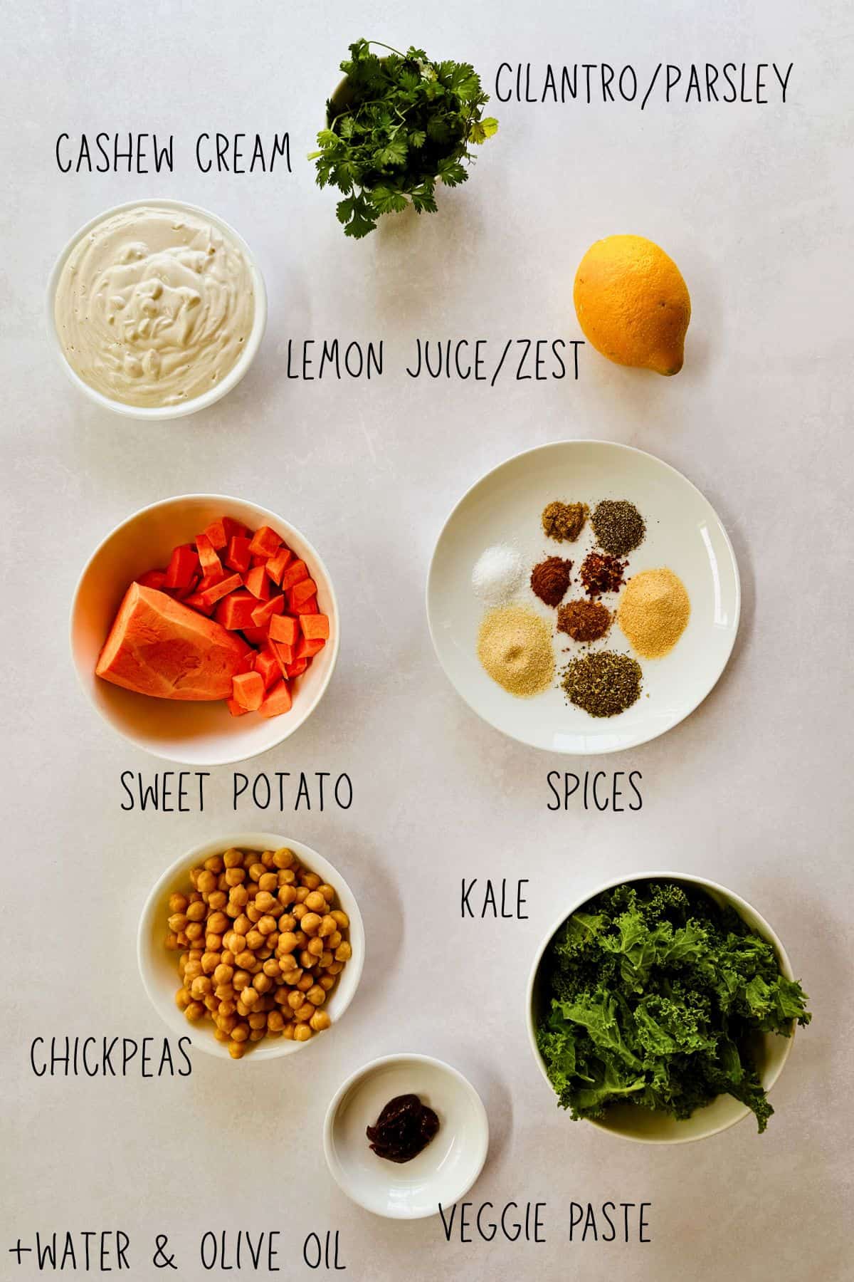 Overhead view of ingredients for creamy chickpea and sweet potatoes arranged on a light surface. Bowls contain chopped sweet potato, kale leaves, chickpeas, spices, and cashew cream. A lemon, fresh cilantro or parsley, and a small dish of veggie paste are also shown. Text labels identify each ingredient, including water and olive oil.