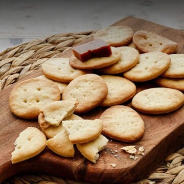 Golden Cuban crackers (Galletas Cubanas) on a wooden board, some broken to show their crisp texture, with a piece of guava paste on top of one cracker.
