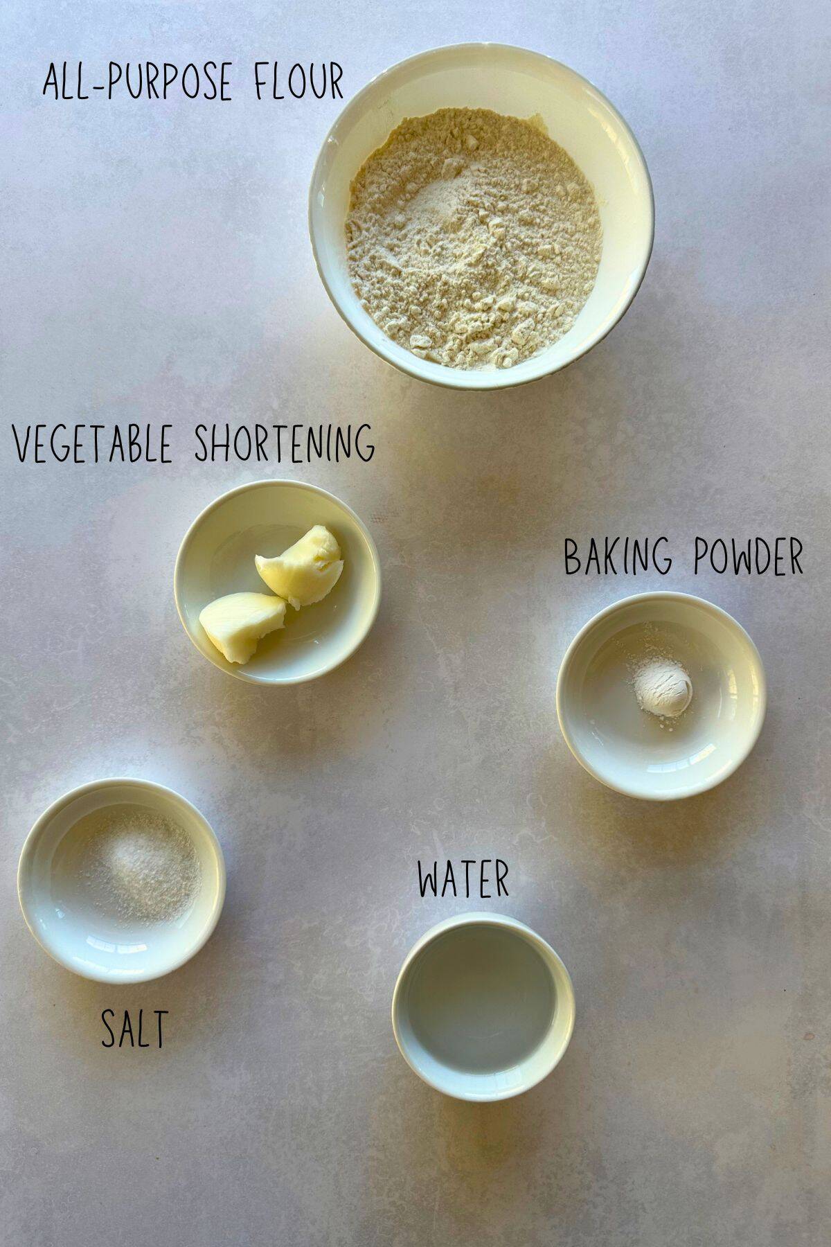 Overhead view of ingredients for Cuban Crackers: bowls of flour, baking powder, salt, vegetable shortening, and water labeled on a light surface.