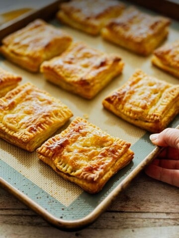 Golden, flaky pastelitos de guayaba fresh from the oven on a silicone-lined tray, glossy and warm, as a hand lifts the edge to serve.