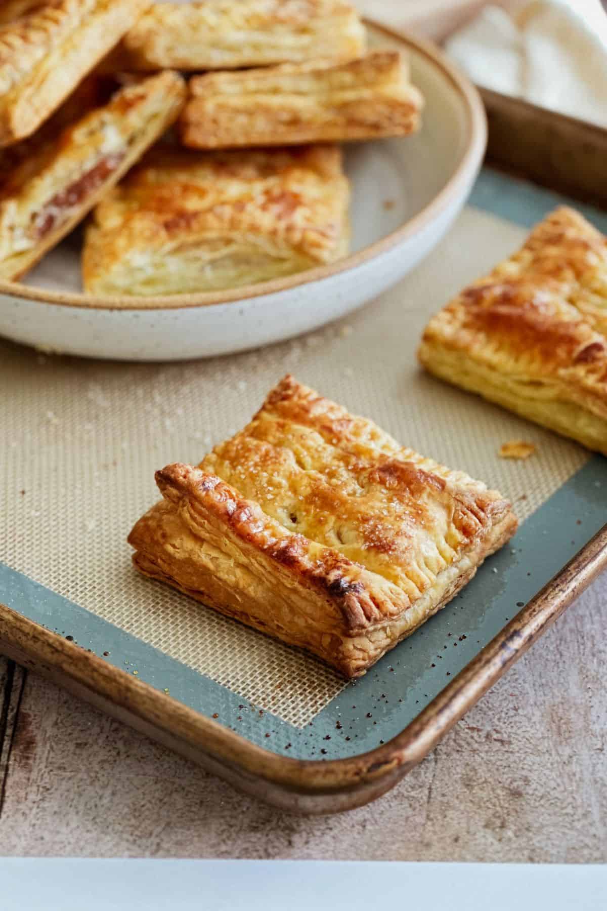 A close-up of freshly baked pastelitos de guayaba on a baking tray. One square pastry is in focus, showing its crisp, flaky layers and caramelized edges, with a plate of more pastries blurred in the background.