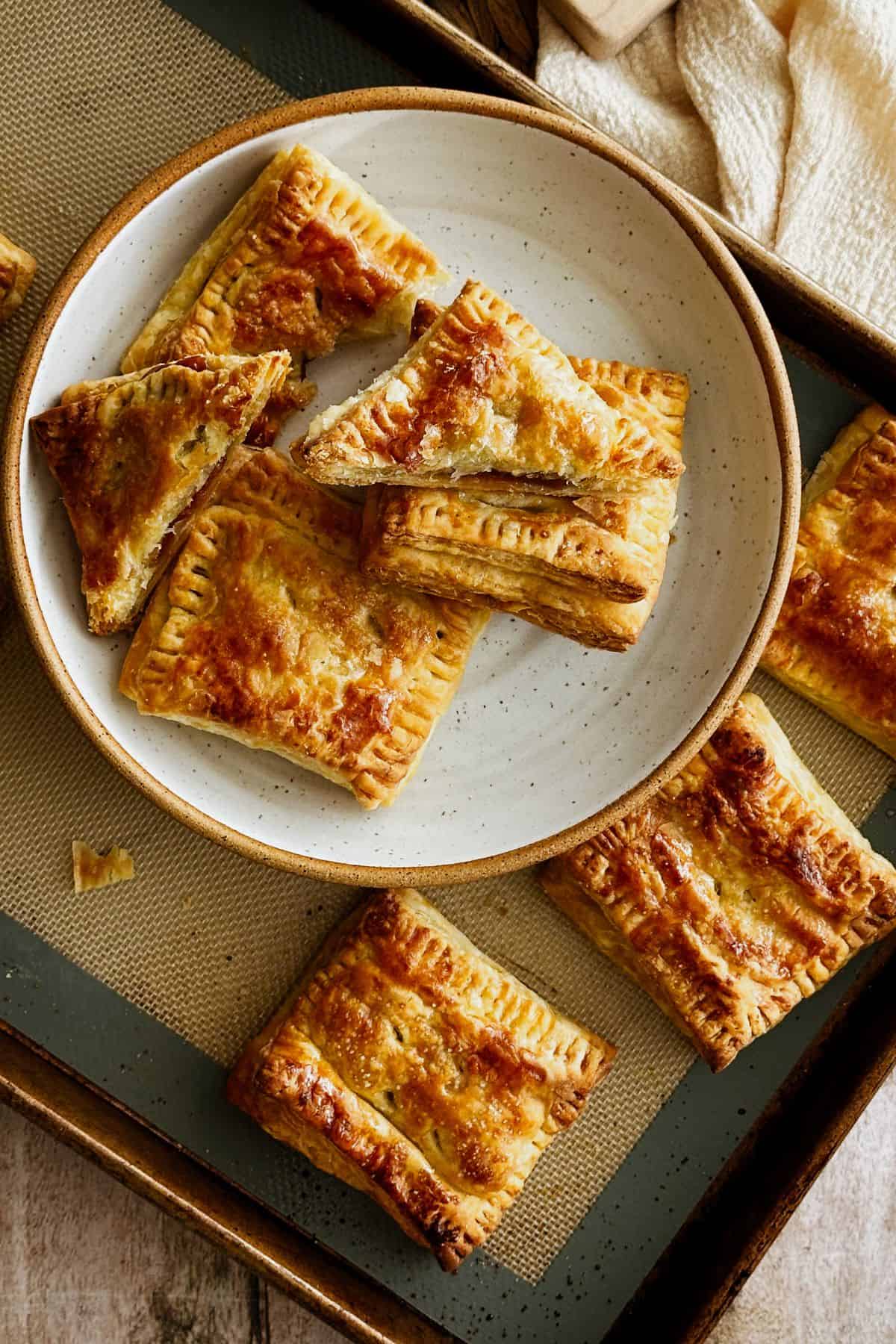 A plate filled with golden, flaky pastelitos de guayaba, some whole and a few cut in half to show the gooey guava filling inside. The plate sits on a baking tray with more pastries around it, all perfectly browned and glossy from the syrup glaze.