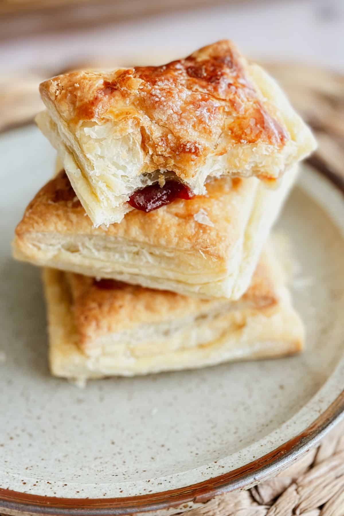A stack of golden, flaky pastelitos de guayaba on a ceramic plate. The top pastry has a bite taken out, showing the gooey red guava filling peeking through the crisp, buttery layers. The focus highlights the delicate texture and caramelized top of the pastry.