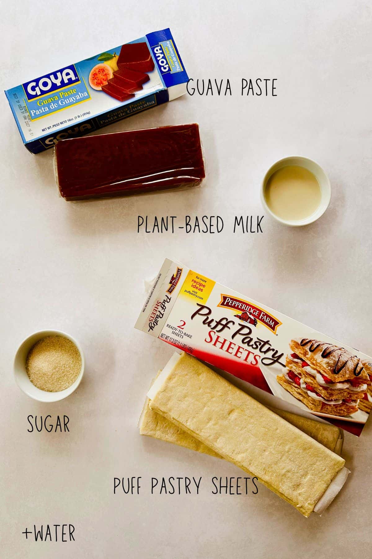 A flat lay of ingredients for making pastelitos de guayaba. There's a package of Goya guava paste with the block of guava unwrapped in front, a small bowl of plant-based milk, a small bowl of sugar, and a box of Pepperidge Farm puff pastry sheets with the dough visible. Text labels point to each ingredient: guava paste, plant-based milk, sugar, puff pastry sheets, and water.