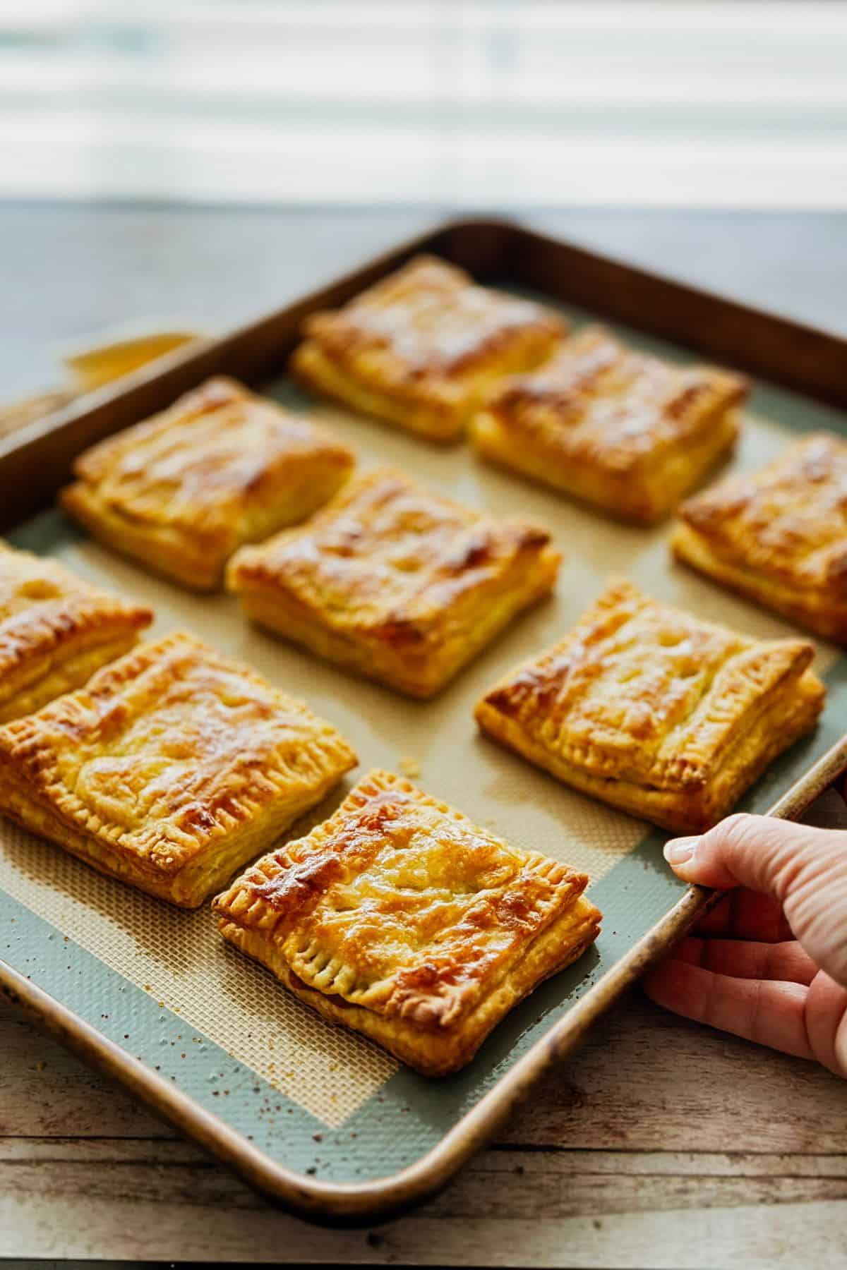 A tray of freshly baked pastelitos de guayaba coming out of the oven. The pastries are golden brown, flaky, and slightly glossy on top, arranged neatly on a baking sheet lined with a silicone mat. A hand is holding the edge of the tray, ready to serve.